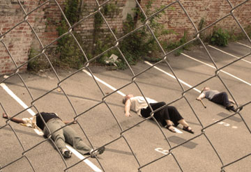 Carrie, Petra and Anita lying in the parking demarkations in a car park, the photo is shot through a wire fence